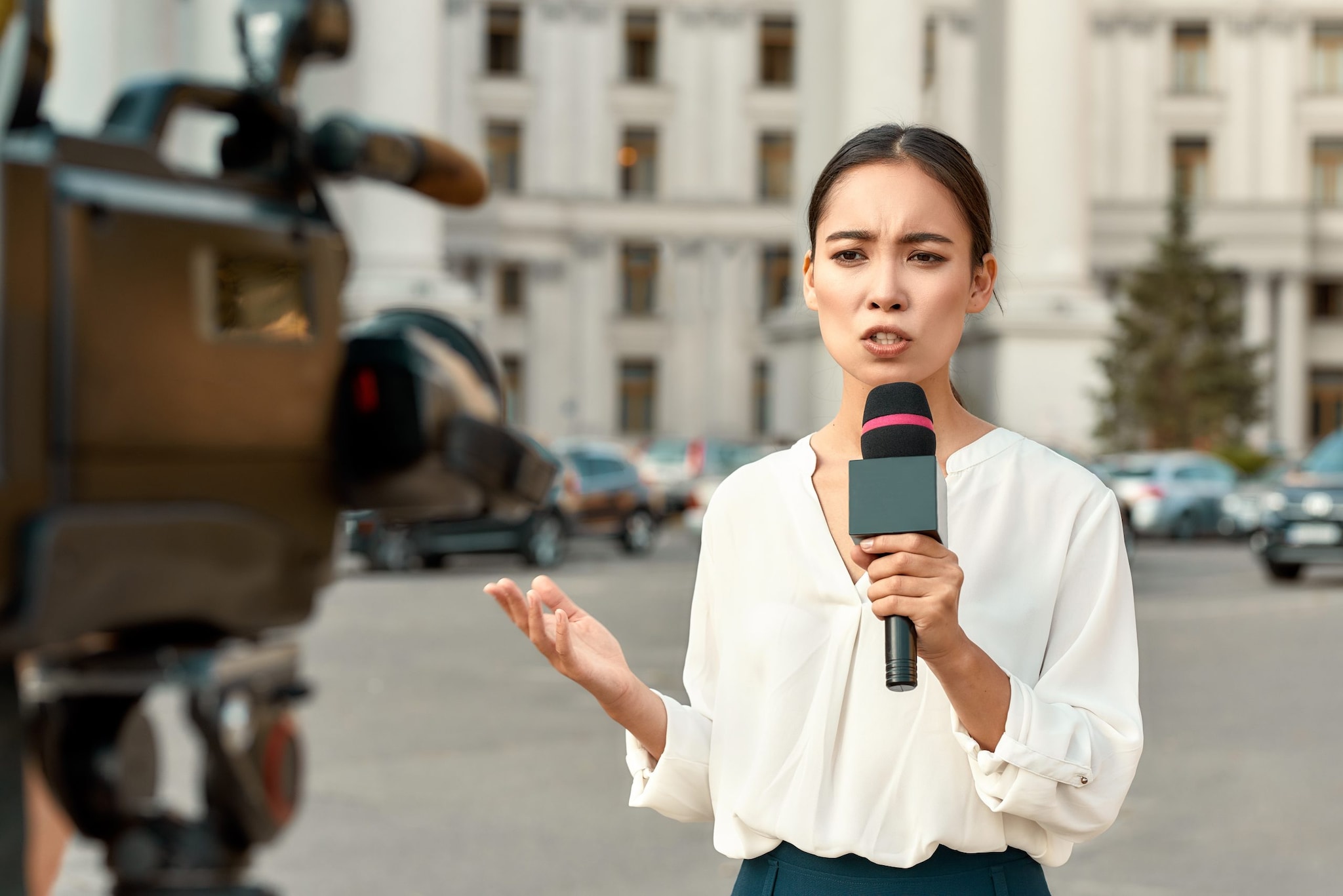 A female reporter is speaking into a camera while holding a microphone during a live steam on a street