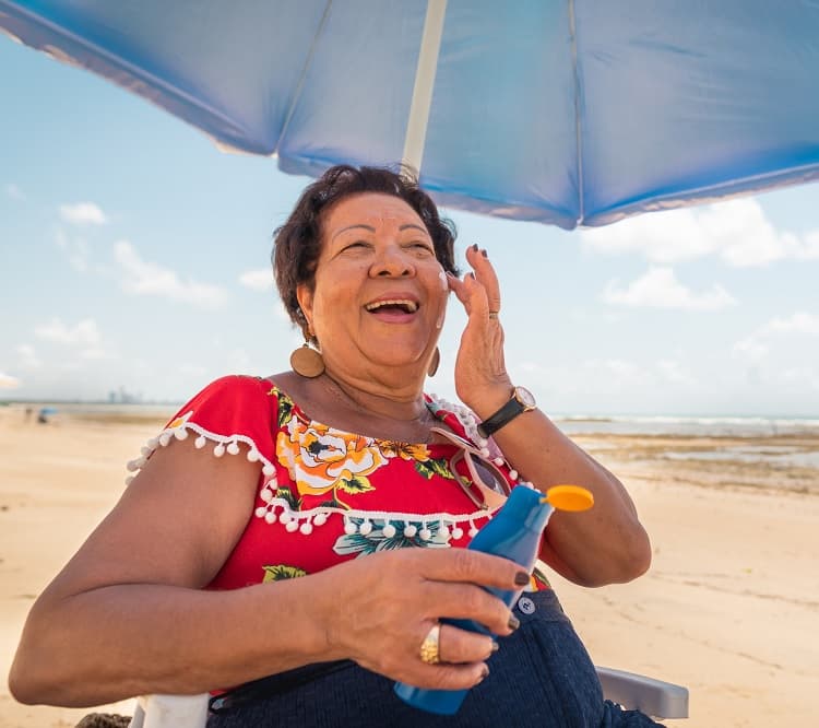 Photo of an older woman sitting under an umbrella on the beach, applying sunscreen