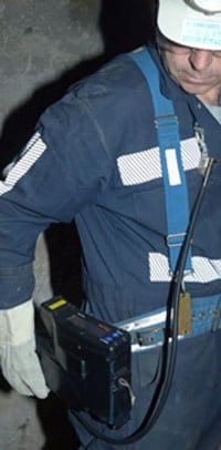 “Minero usando el monitor personal continuo de polvo PDM3700. El aire entra por una boquilla que se coloca en la solapa del minero y llega a través de un tubo al dispositivo que se sujeta a la altura de la cadera en su cinturón. El minero está mirando hacia abajo para leer la pantalla digital”.