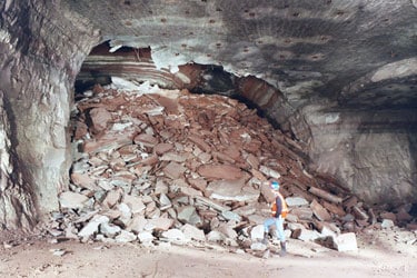 Fotografía de un minero observando el derrumbe del techo de una mina subterránea. El derrumbe causó una acumulación de roca de quince pies de altura por veinte de ancho (o 4.5 metros de altura por 6 de ancho) que bloquea la entrada completamente. 