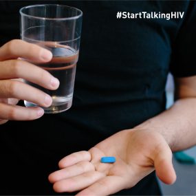 Image displays the upper body of a man who is holding a glass of water in one hand and a blue pill in the palm of his other hand.