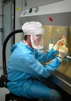 Man is wearing a powered air purifying respirator, gloves, and solid-front gown. He is working within a BSC, and self-closing, locked doors are in the background.