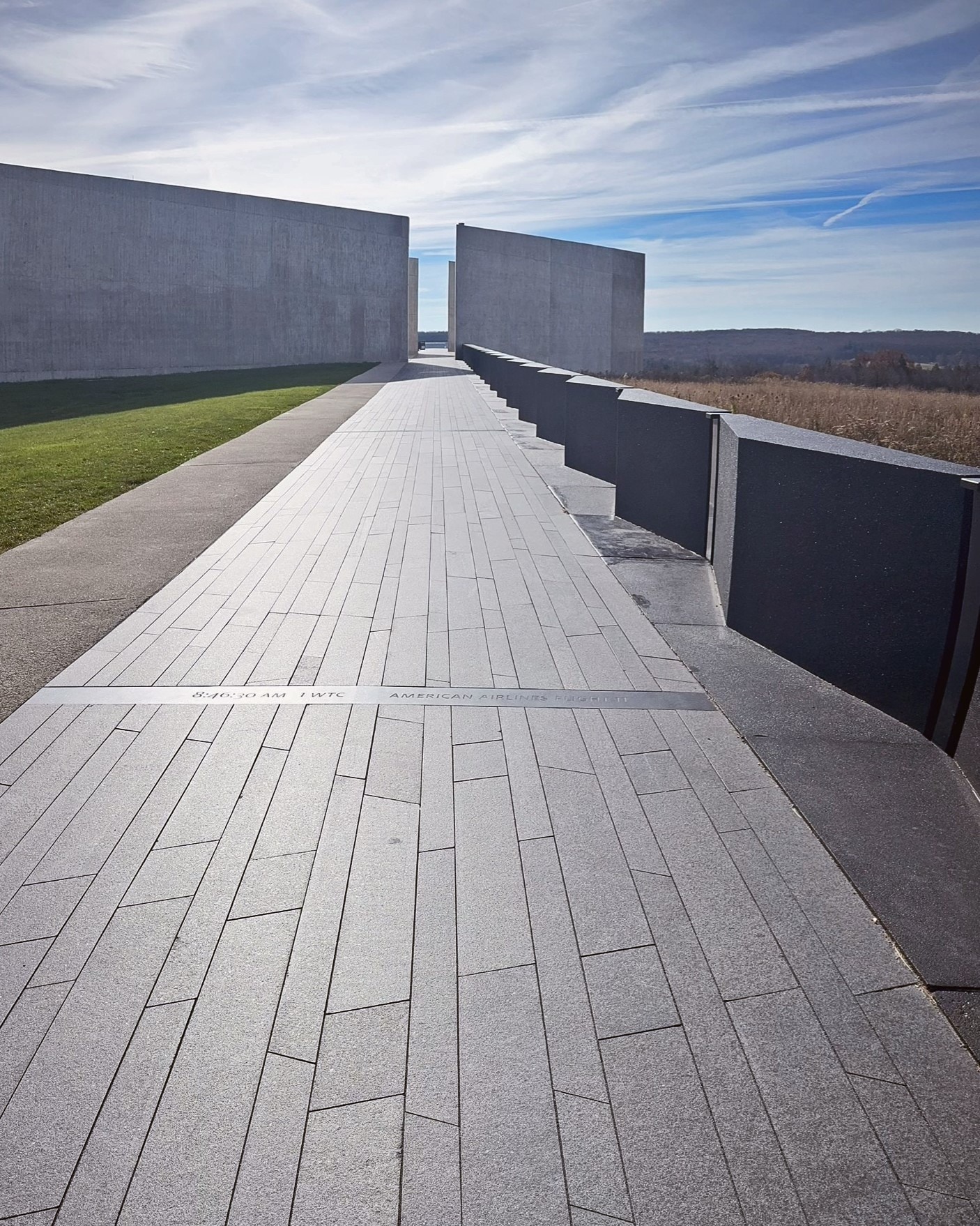 A black granite walkway, situated at the Visitor Center of the Flight 93 Memorial , guides visitors through the Portal Walls, tracing a solemn journey along the Flight 93 flight path. The glow of the bright sun in the top left illuminates the path, symbolizing hope and resilience.