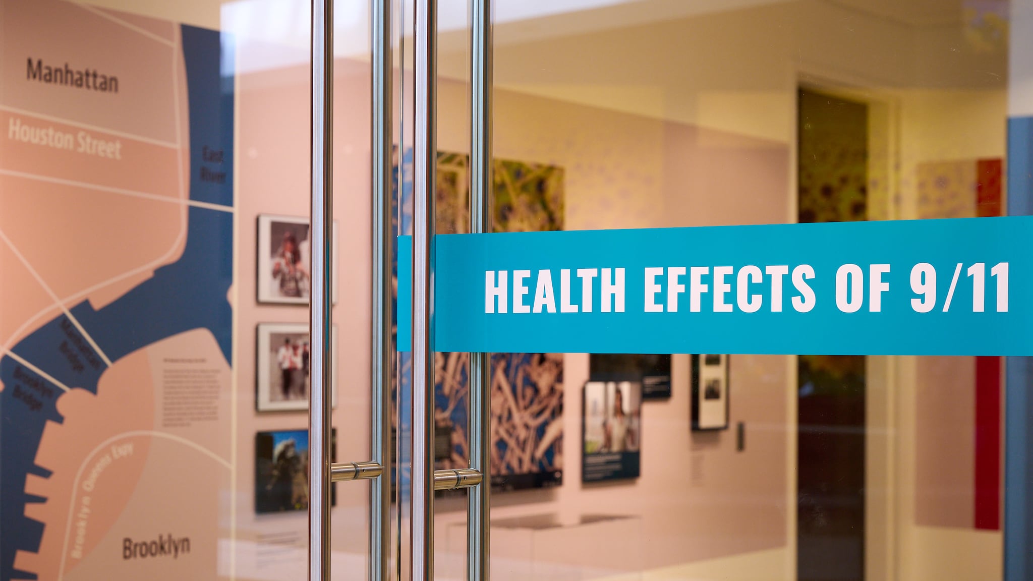 A captivating view through a glass door adorned with the exhibition title at the Health Effects of 9/11 exhibit. Beyond the door, glimpses of the exhibit come into focus, offering a symbolic look into the shared experiences and remembrance encapsulated within the display.