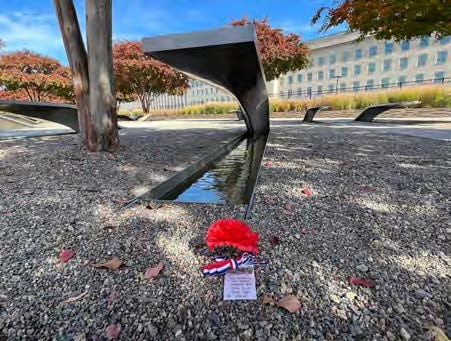 A steel cantilevered bench over a pool of water, engraved with the name of a Pentagon 9/11 attack victim serves a focal point. A red flower adorned with a red, white and blue ribbon is delicately in front of the bench. A heartfelt note on the flower pays tribute, creating a scene of remembrance and honor. In the background, a serene setting of trees, additional benches.