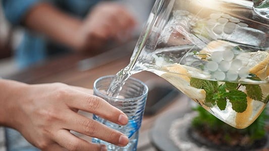A hand pouring water from a pitcher with fruit and mint into a cup.