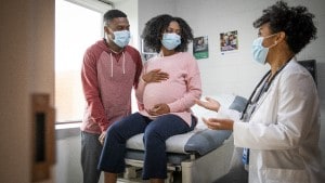 Man with pregnant woman wearing masks and talking with a masked doctor in exam room