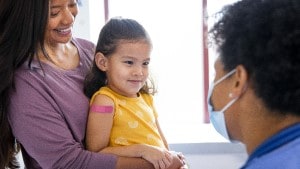 Mom holding preschool-age girl with Band-Aid on her arm as they talk to doctor in exam room