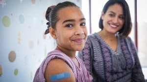 Girl looking at camera smiling with Band-Aid on her arm as mom looks at her