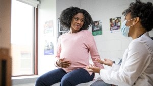 Pregnant Black woman sitting in exam room talking with doctor who is weaking a mask