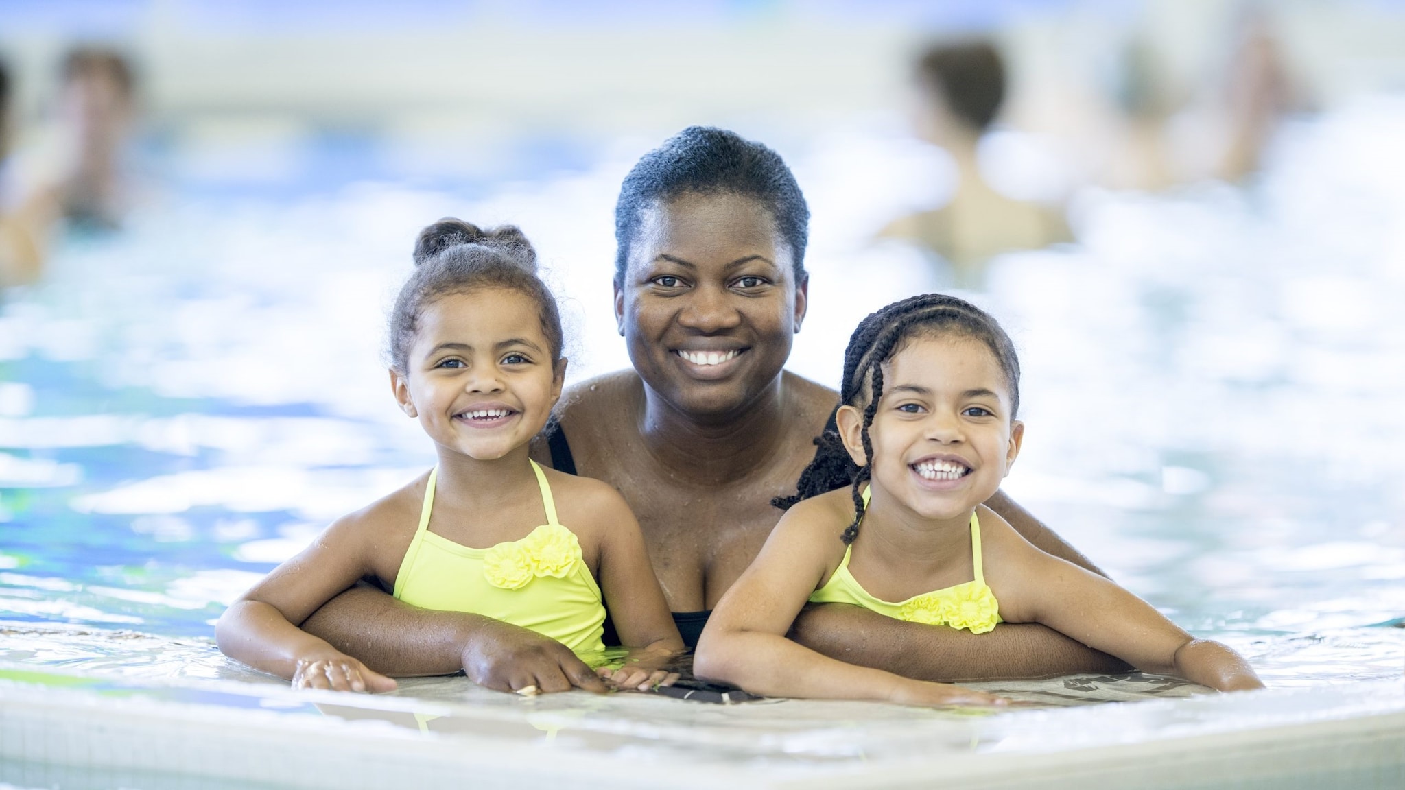 Mom in the pool with her arms around her young daughters who are on either side of her.