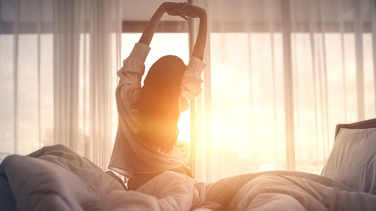 Young woman silhouetted, facing away, stretching sitting in bed.
