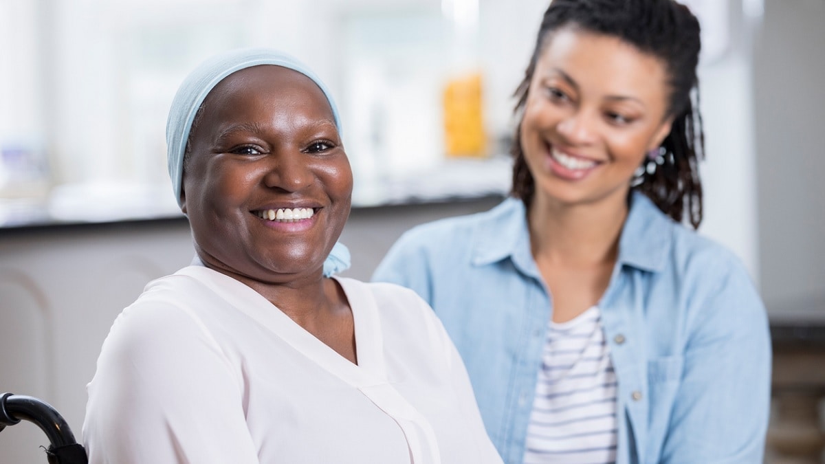 An older African American cancer patient smiles as her adult daughter looks on.