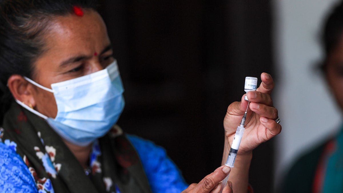 A woman wearing a surgical mask draws liquid from a vial into a syringe.