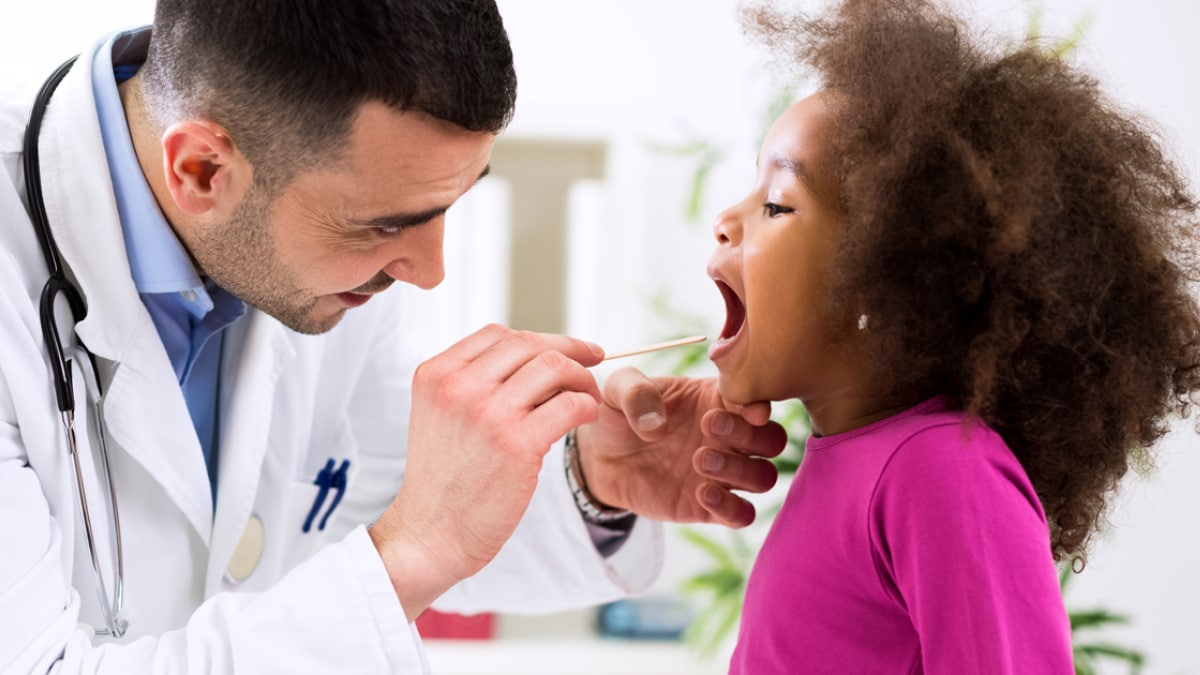 A male doctor examines a little girl's throat using a tongue depressor.