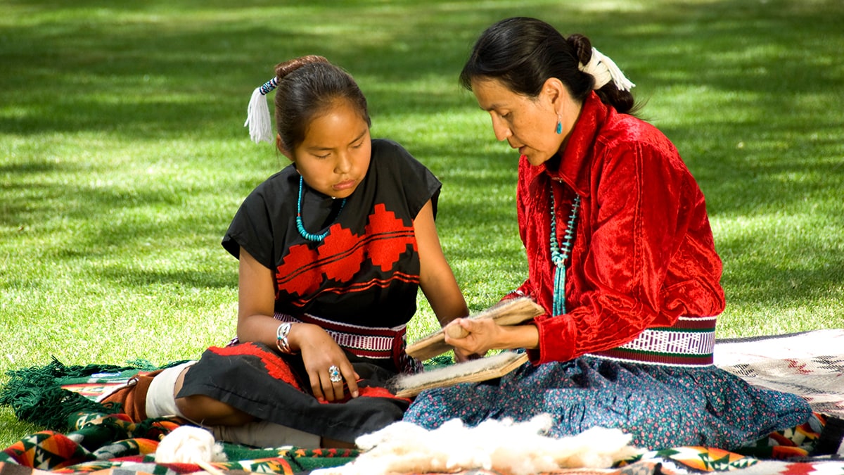Two Native Americans sitting on a blanket with the older one teaching the younger one how to weave