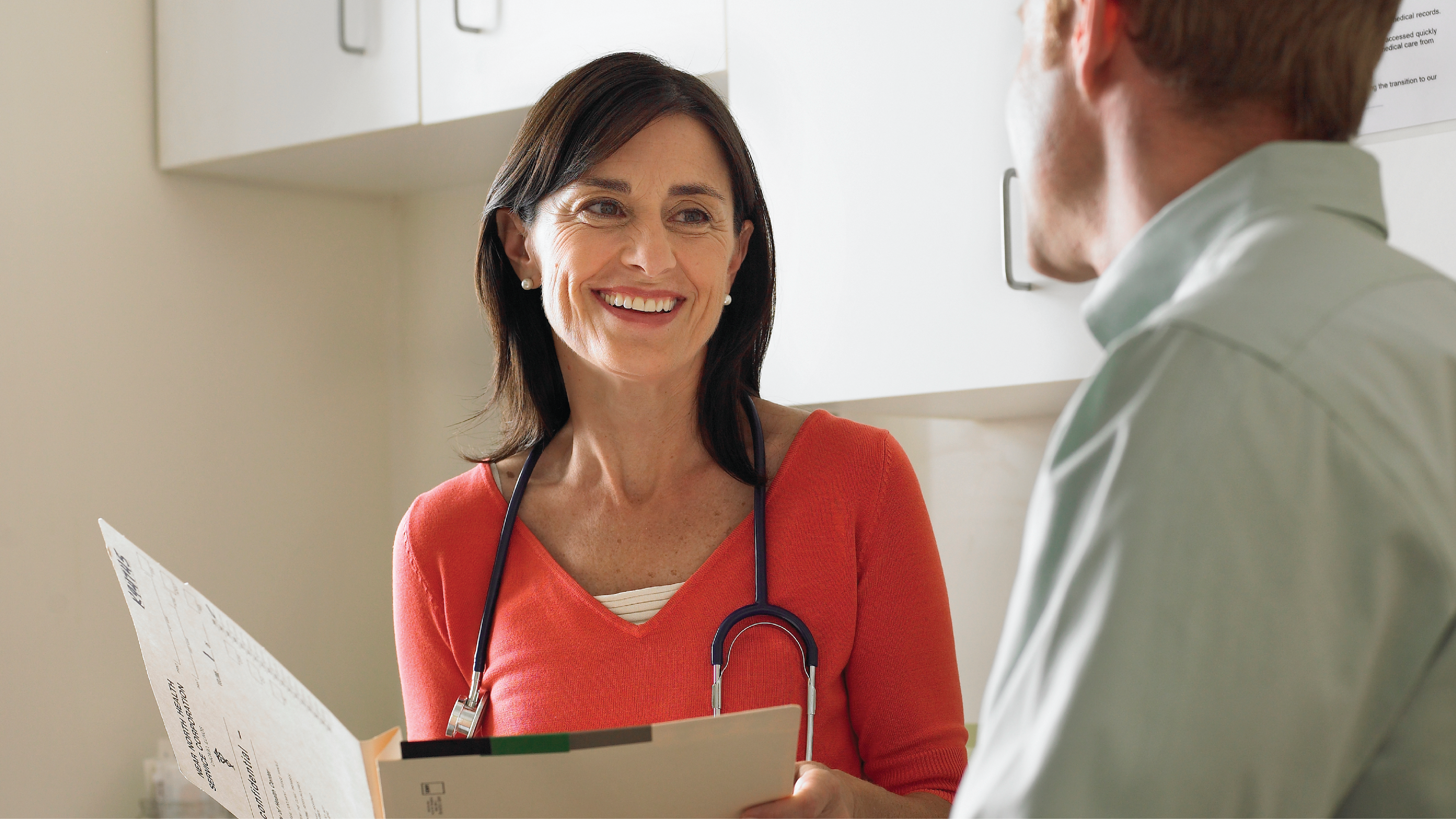 A health care provider smiling and holding a medical chart while talking to a patient.