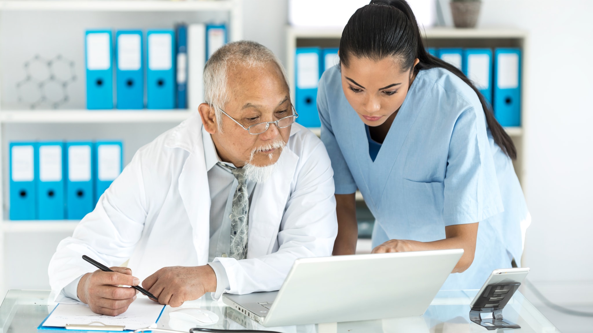 A picture of an older man and a younger woman, both in clinical setting clothing, looking at a laptop computer.