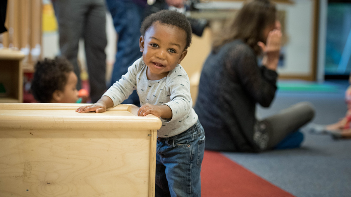 1 year old stands holding onto a wood block