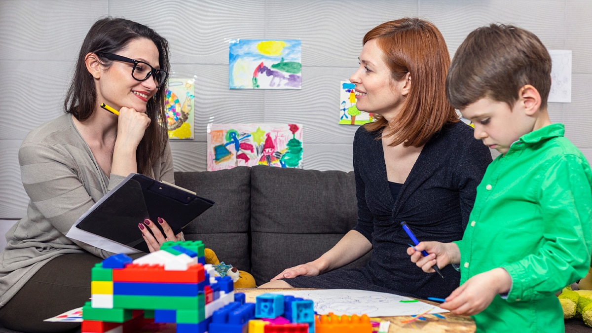 A therapist wearing glasses holds a tablet and smiles during meeting with a mother and her child. There are toy blocks on the table.