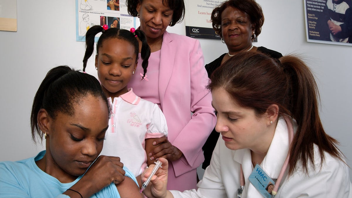 A nurse in the process of administering an intramuscular vaccination into a girl’s left arm whose sister, mother, and grandmother watched from the background