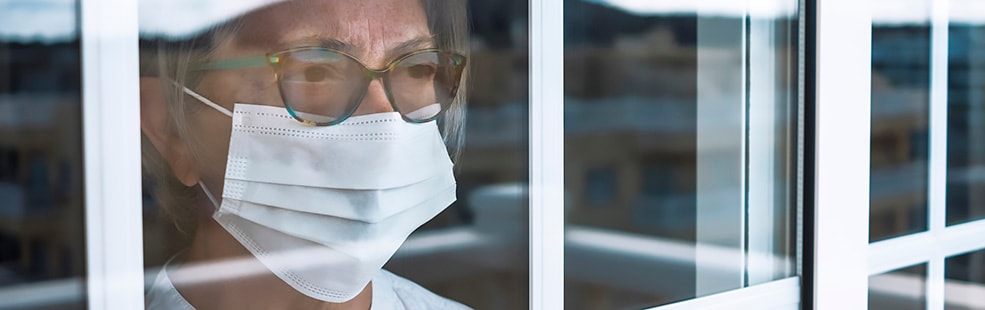 Older woman wearing mask behind the glass of a window in isolation with COVID-19.