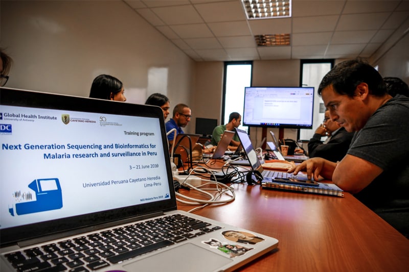 Closeup of laptop with words Next Generation Sequencing and Bioinformatics for Malaria research and surveillance in Peru with attendees in the background