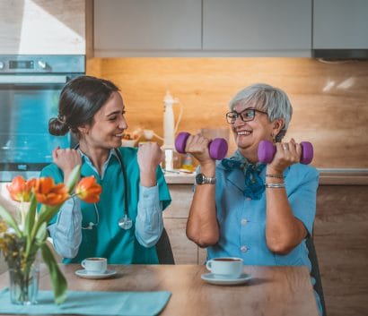 A nurse helping a woman lifting a dumbbell at a table and smiling.