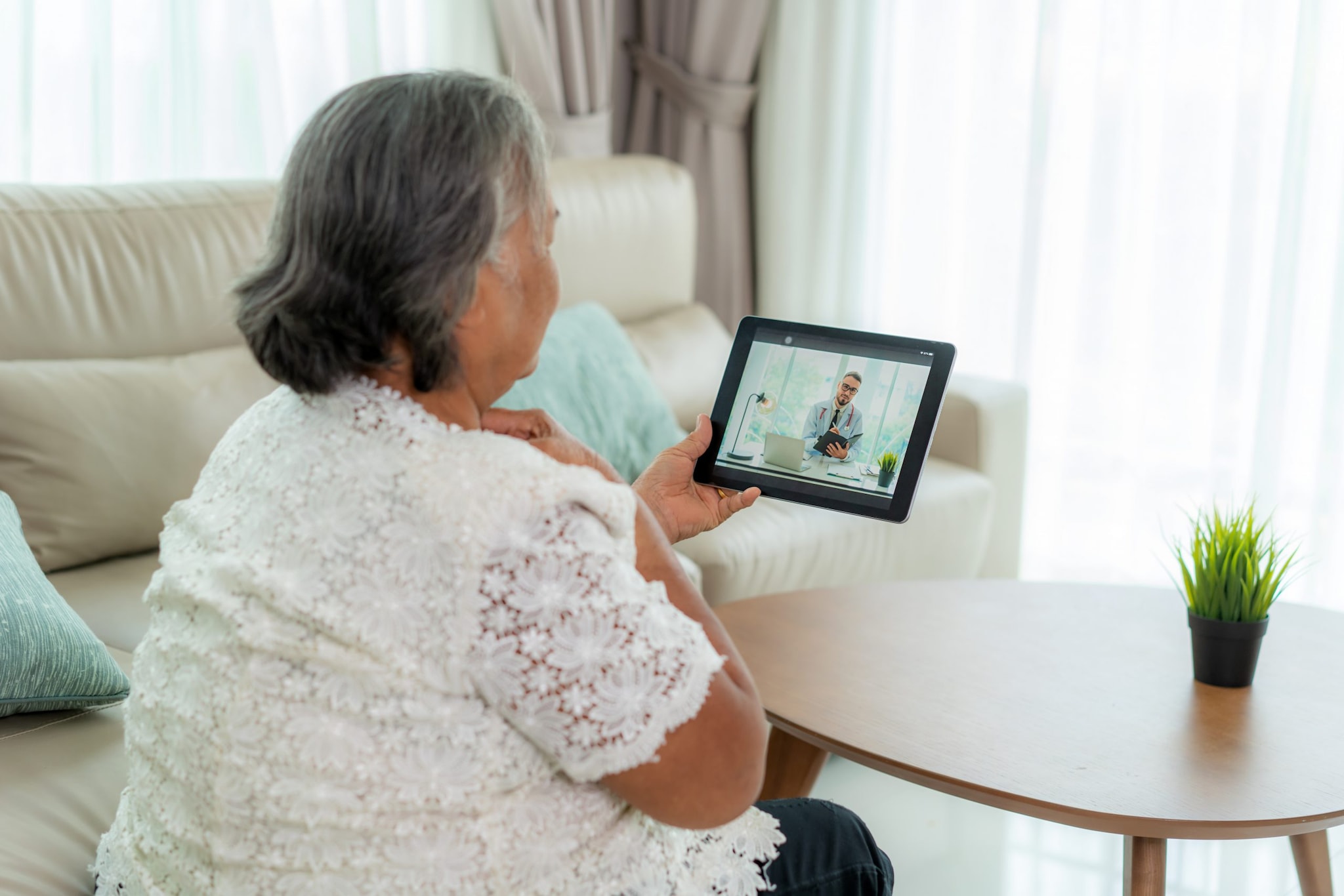 Back view of elderly woman making video call with her doctor with her feeling sore throat on digital tablet online healthcare digital technology service consultation while staying at home.