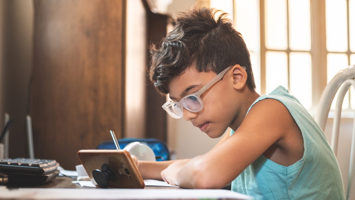 Young boy with glasses taking notes and sitting at desk at home while watching class virtually on electronic screen