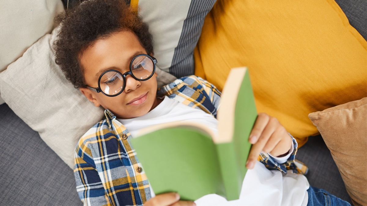 A boy wearing glasses reading a book while lying down on the couch
