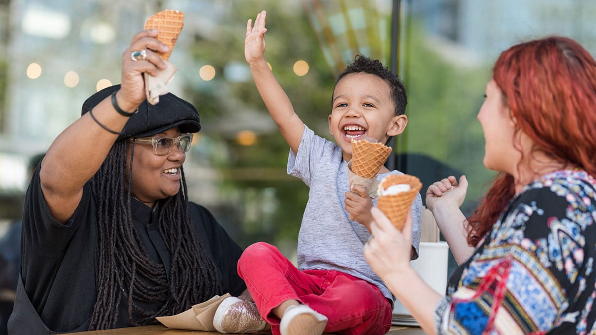 Young child joyfully eating ice cream with two mothers.