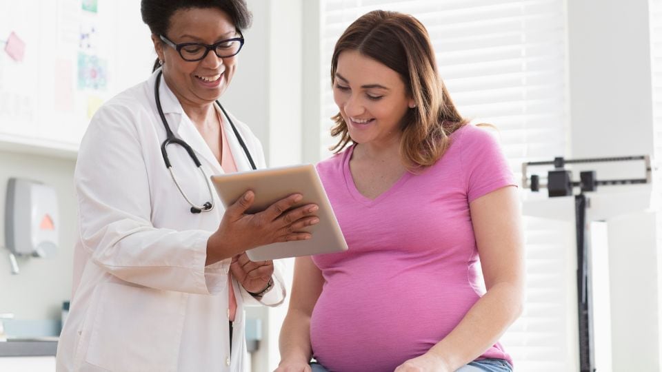 Pregnant person and doctor smiling, looking at a tablet in a healthcare office.