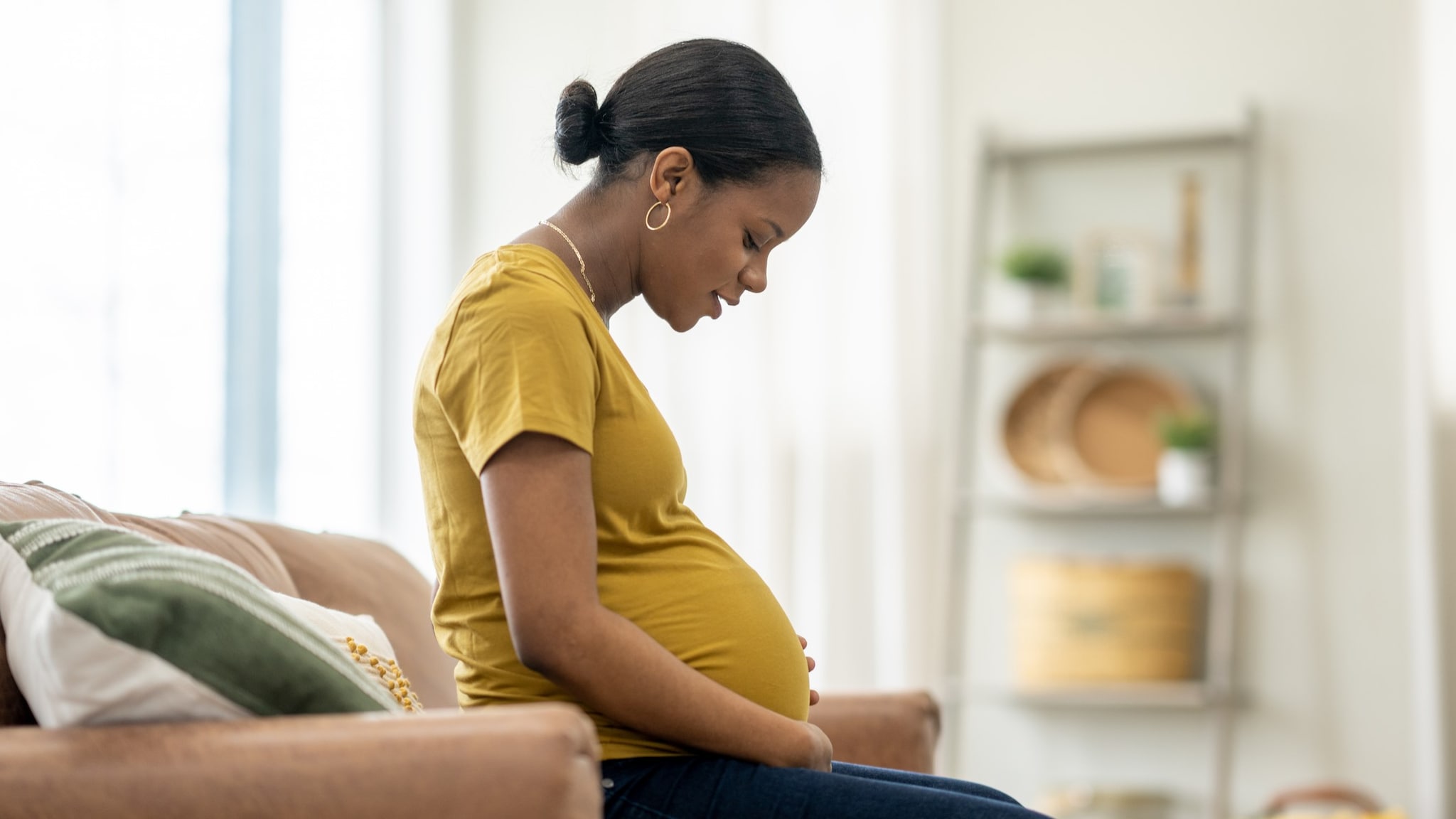 Pregnant person in a yellow shirt sitting on a couch looking at their stomach.