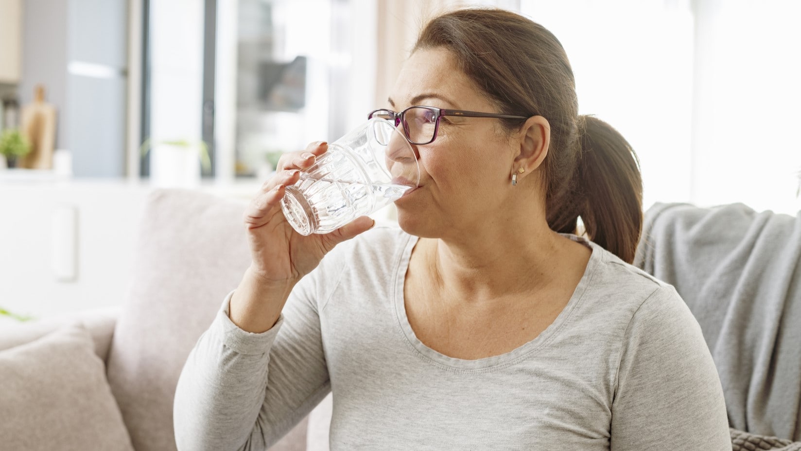 An older adult sitting on a couch drinking a glass of water.