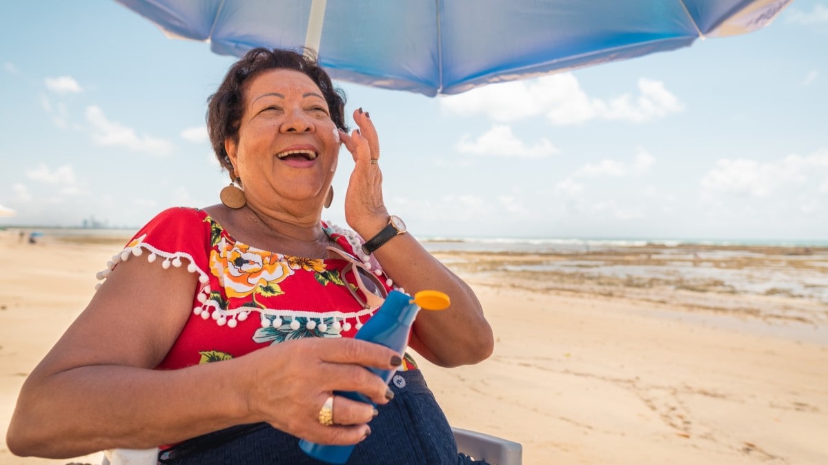 una mujer mayor sentada debajo de un paraguas en la playa, aplicándose filtro solar