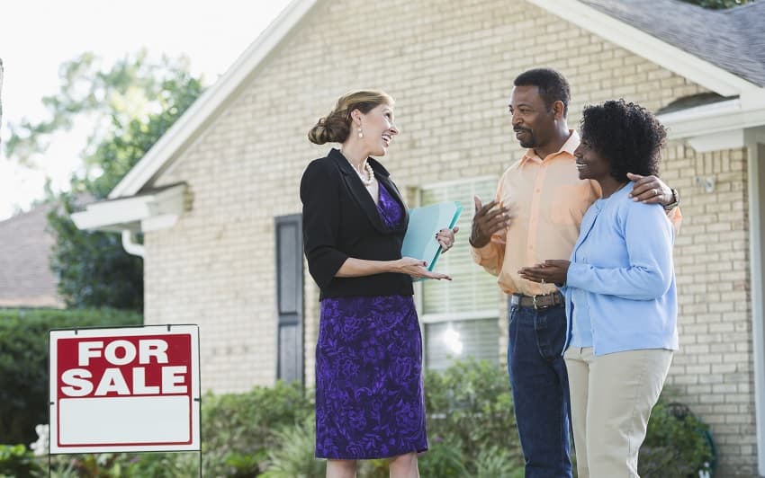 A real estate agent talking to a couple in front of a house with a For Sale sign in the yard
