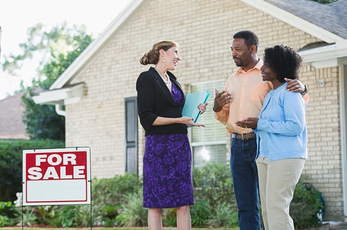 A real estate agent standing in front of a house with a For Sale sign in the yard, talking with a couple who could be the homeowners, or potential buyers.
