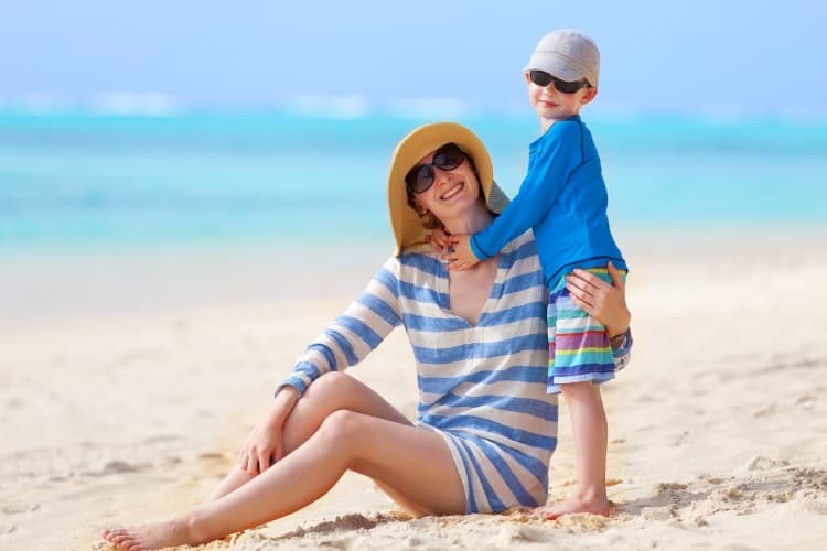 Photo of a mother and child on the beach. Both are wearing sunglasses, hats, and long-sleeved shirts.
