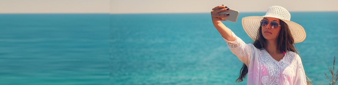 Show Us Your #SunSafeSelfie! Photo of a woman on a beach wearing a hat and sunglasses taking a photo of herself