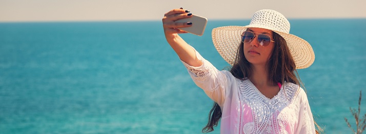 Show Us Your #SunSafeSelfie! Photo of a woman on a beach wearing a hat and sunglasses taking a photo of herself