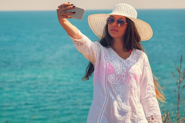 Photo of a woman on the beach wearing a long sleeved shirt, hat, and sunglasses taking a picture of herself