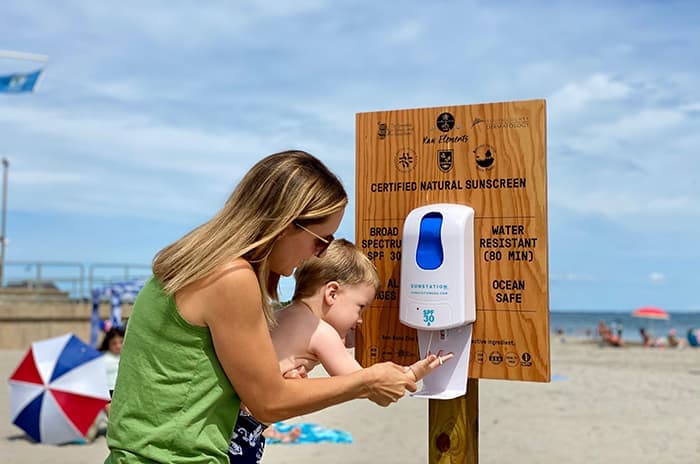 Foto de una mujer que ayuda a su hijo pequeño a aplicarse protector solar de un dispensador en Scarborough State Beach en Rhode Island