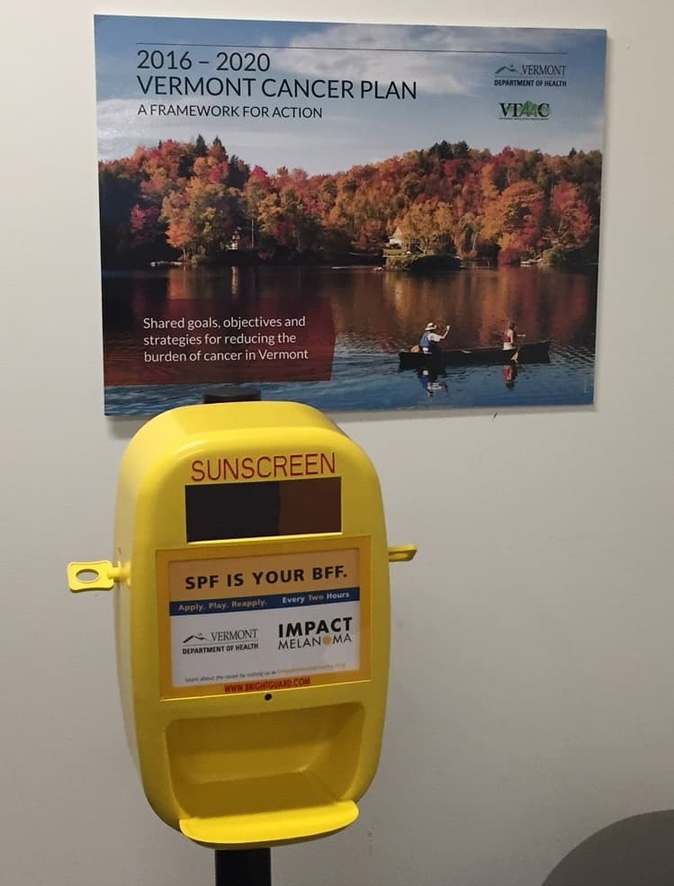 Photo of a sunscreen dispenser in an office underneath a poster promoting the 2016 - 2020 Vermont Cancer Plan.