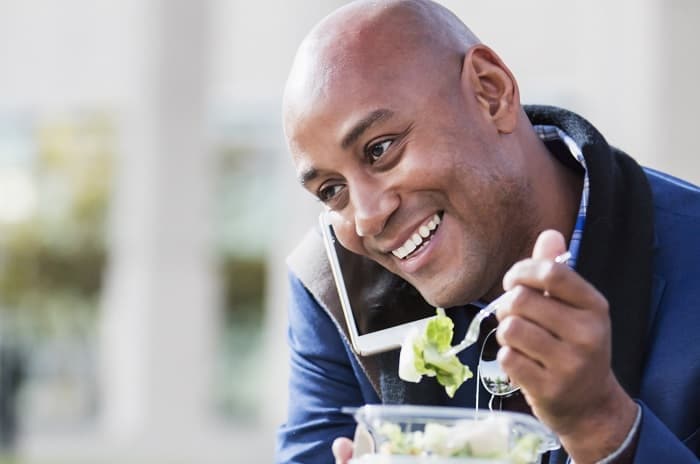 Foto de un hombre comiendo una ensalada mientras está sentado en un banco de un parque hablando en su teléfono celular