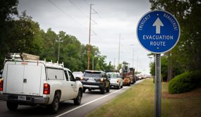 Cars and trucks line up in traffic on a road. In the foreground, a round blue road sign says “Emergency Management Evacuation Route” with an arrow pointing the way the vehicles are going.