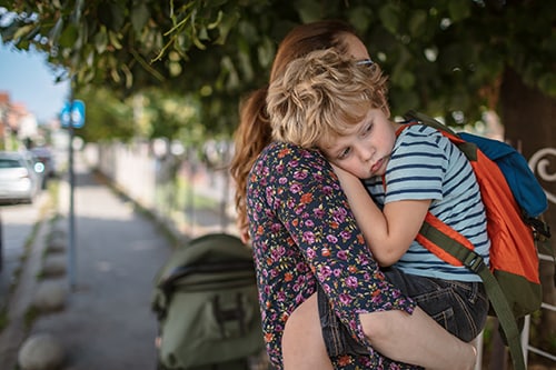 Mother holding unhappy preschooler who is wearing a backpack