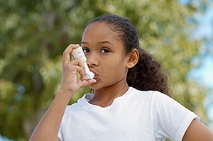 A young girl in a white T-shirt using an inhaler outside.
