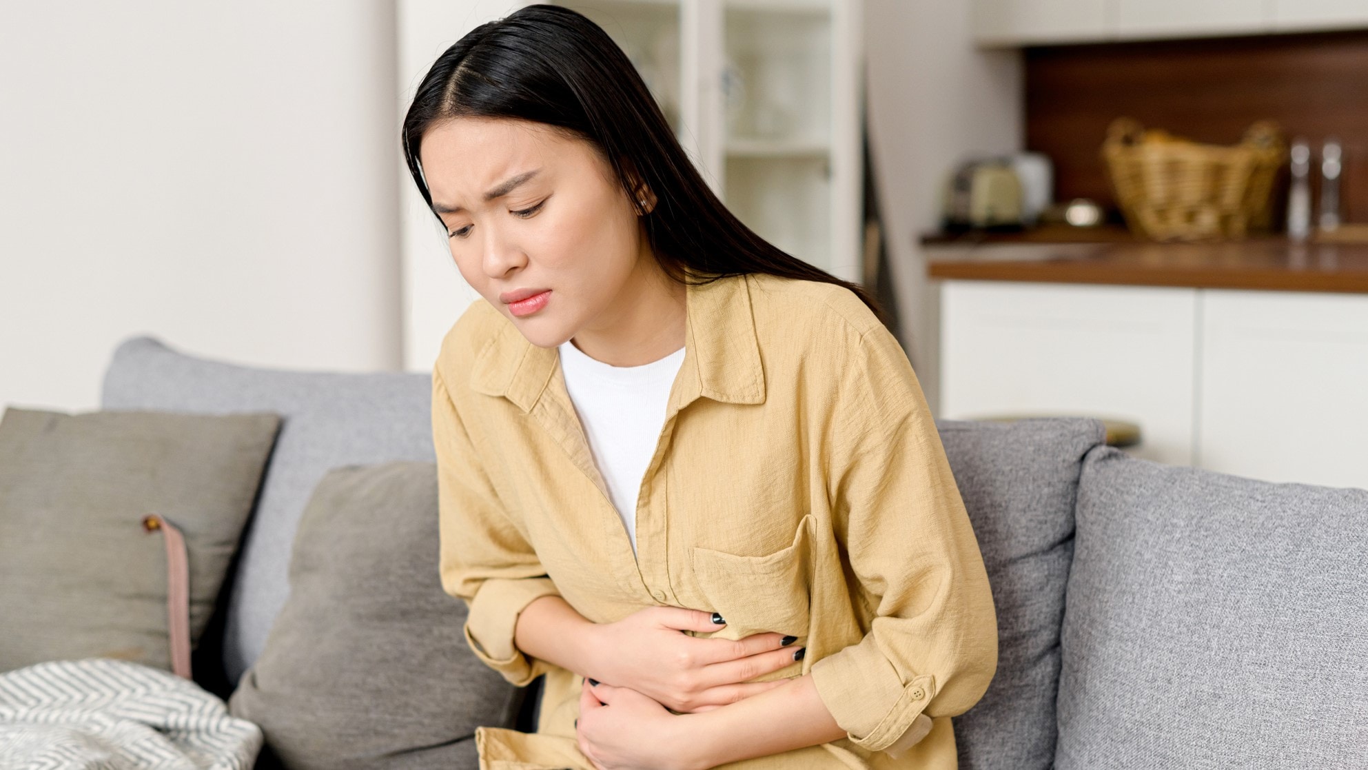A person with long black hair sitting on a couch holding their cramping stomach.