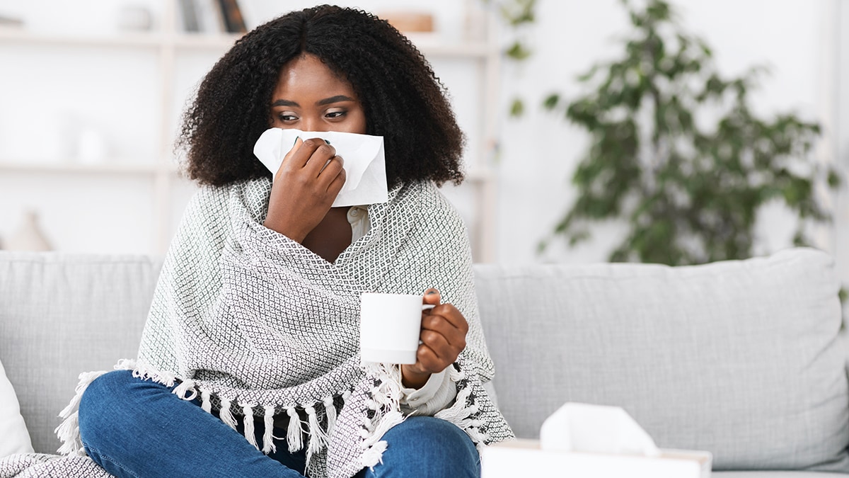 Sick woman on a couch draped in a blanket drinking tea and treating her cold symptoms.
